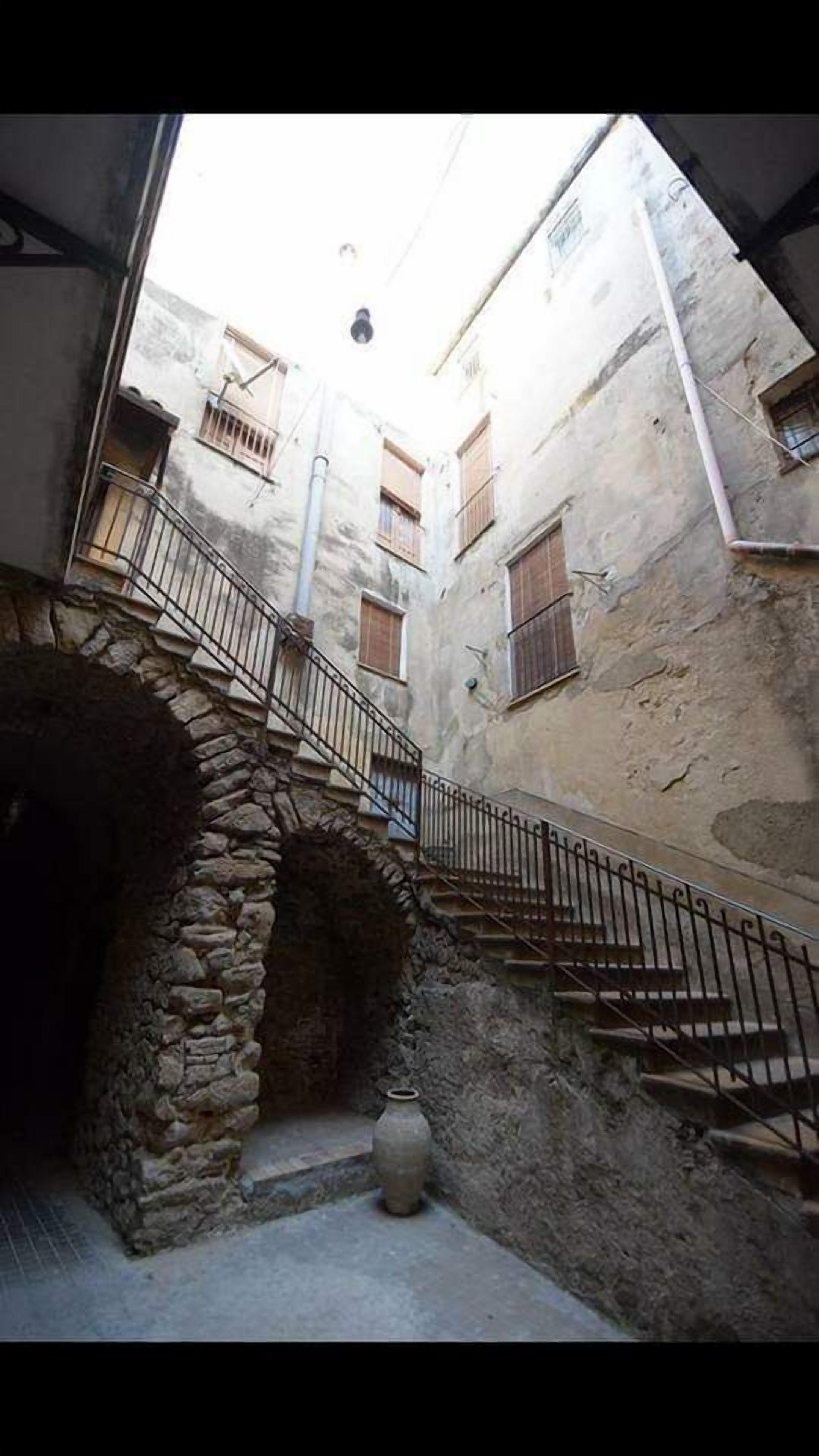Un estrecho callejón con paredes de piedra, una escalera metálica y un jarrón grande al final.