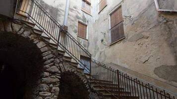 Un estrecho callejón con paredes de piedra, una escalera metálica y un jarrón grande al final.