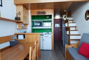 Een kleine keuken met een magnetron, fornuis en spoelbak, een zithoek met houten meubels, en een trap naar een slaapgedeelte met een slaapbank.