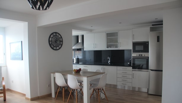 Una cocina moderna con armarios blancos, encimera negra y electrodomésticos de acero inoxidable. Hay una mesa de comedor con cuatro sillas y un frutero encima.