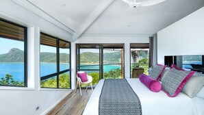 A modern bedroom with a large bed, a ceiling fan, and a view of the sea.