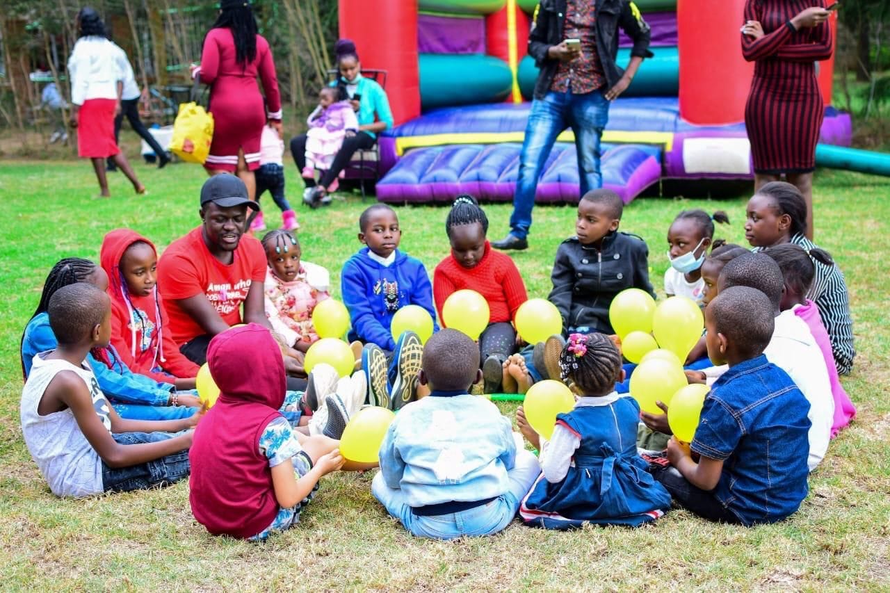 Niños sentados sobre la hierba con globos, un castillo inflable y adultos al fondo.