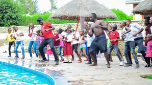 A group of people dancing outdoors near a pool with thatched-roof structures in the background. - Naivasha Peppercorn Holiday Resort (Naivasha)