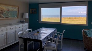 A dining area with a table and chairs, a view of the sea, and a painting on the wall. - Maison vue mer côte sauvage pointe de la Torche (Penmarch)