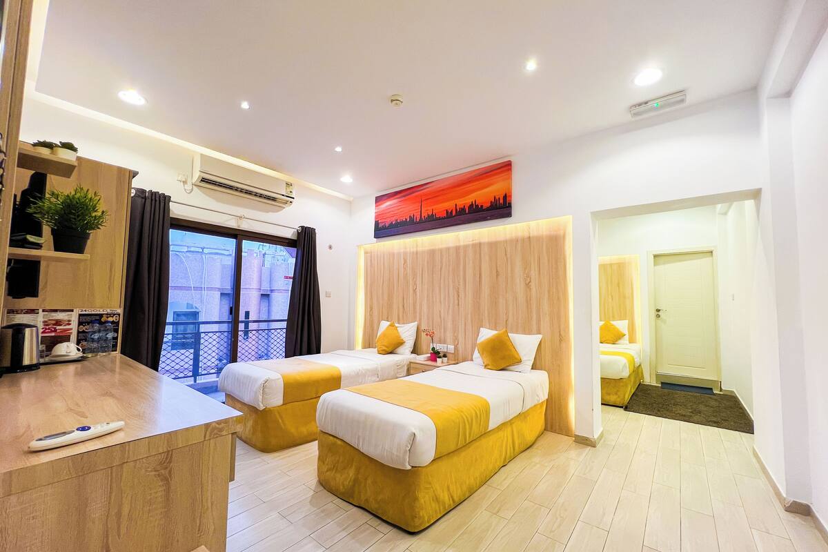 Een hotelkamer met twee bedden, een bureau, een raam met gordijnen, een airconditioning en een stadsgezichtschilderij aan de muur.