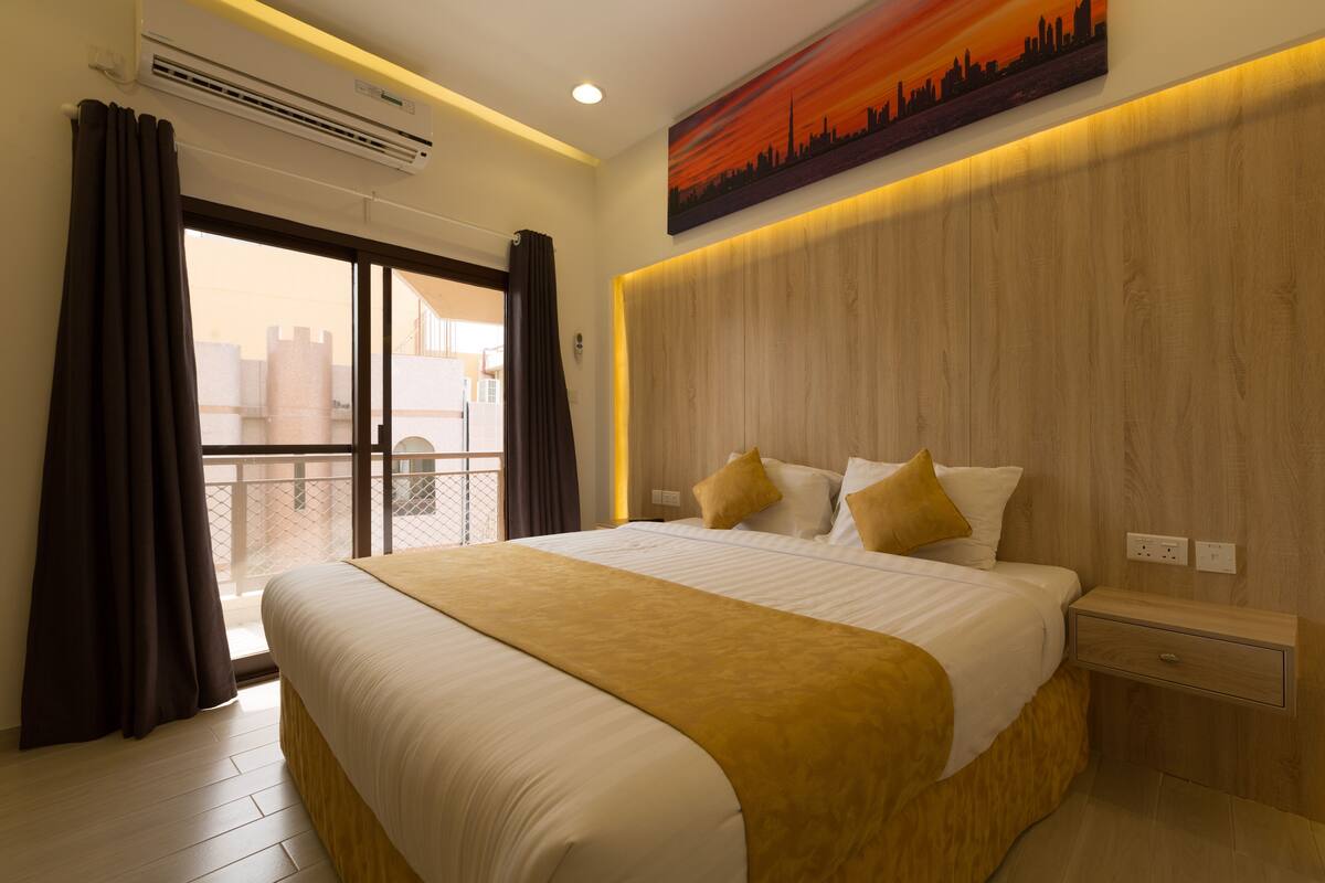 Een hotelkamer met een groot bed, een houten hoofdeinde, een nachtkastje en uitzicht op de stad vanaf het balkon.
