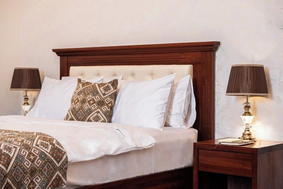 Un letto rifatto con cura, un copriletto decorato, due abat-jour e un comodino in legno con un libro e una lampada.