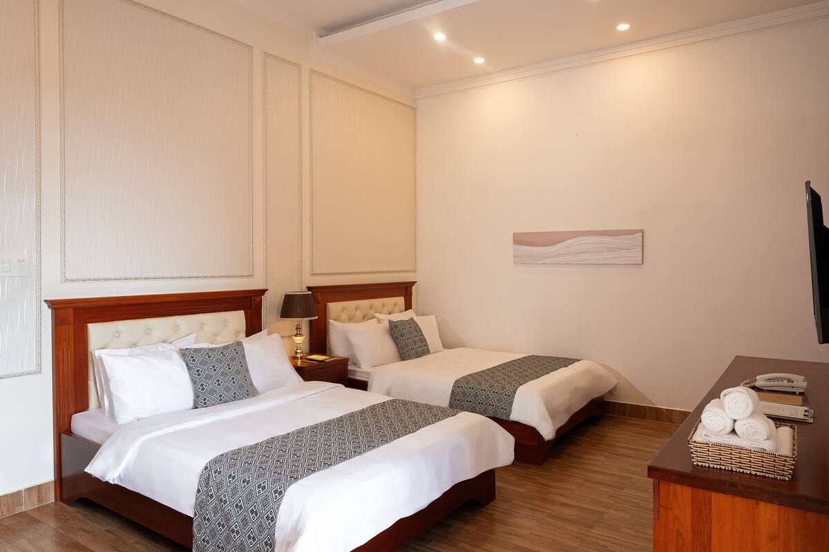 Une chambre d’hôtel avec deux lits, une tête de lit en bois, une table de chevet avec une lampe, un petit bureau avec un téléphone et un panier rempli de serviettes.