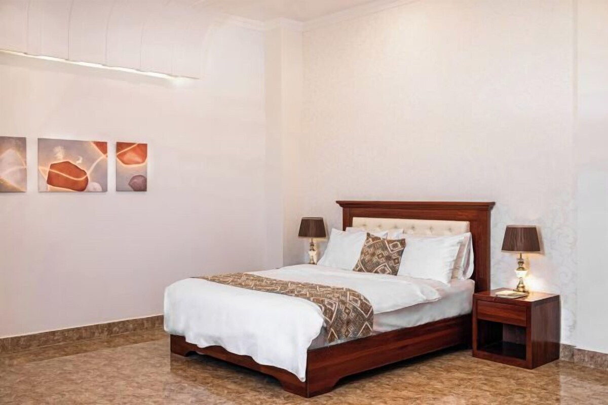 Een keurig opgeruimde slaapkamer met een houten bed, twee nachtkastjes met lampen en drie kunstwerken aan de muur.