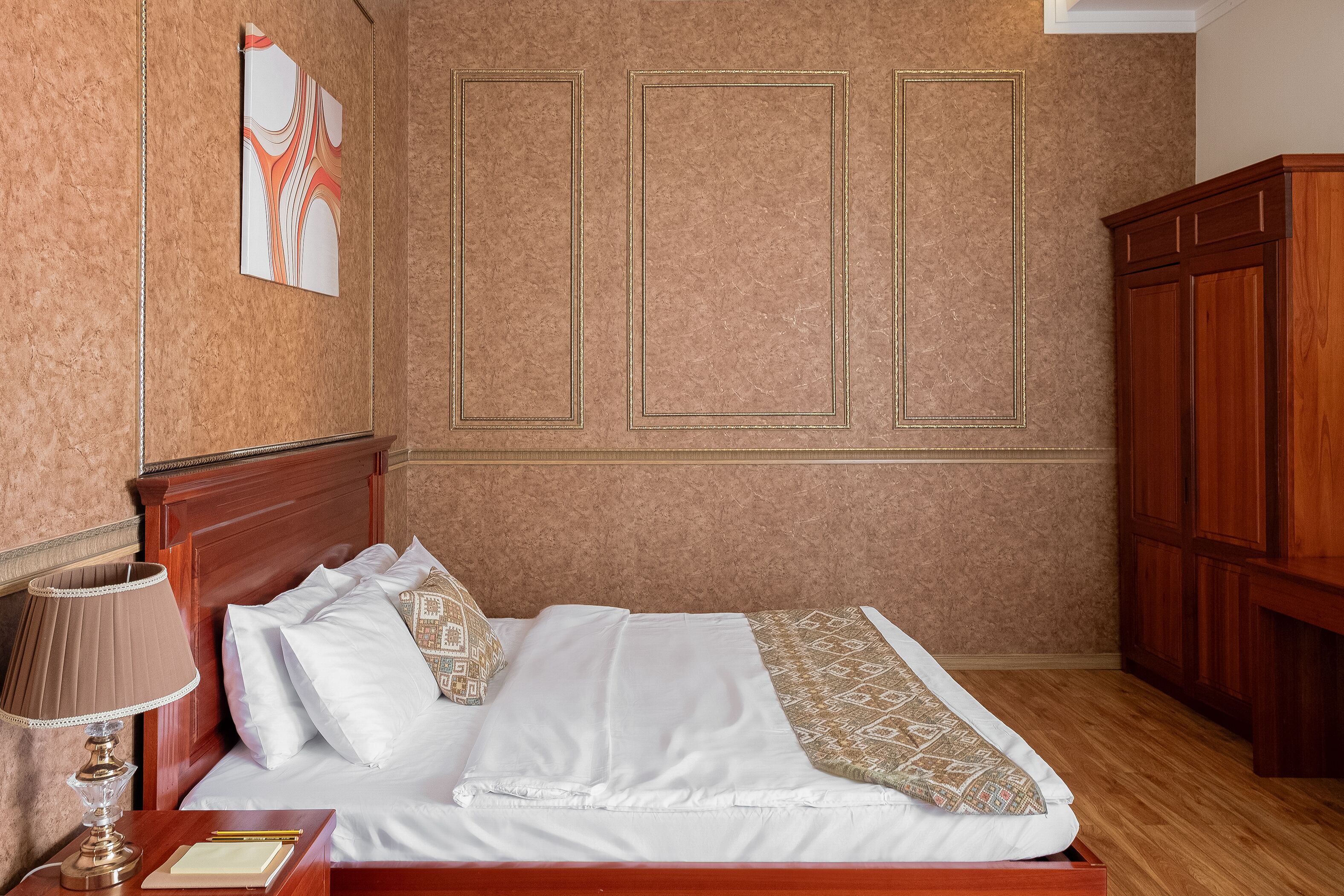 Habitación de hotel con cama de madera, mesita de noche con lámpara, armario y una pared decorada con un cuadro.