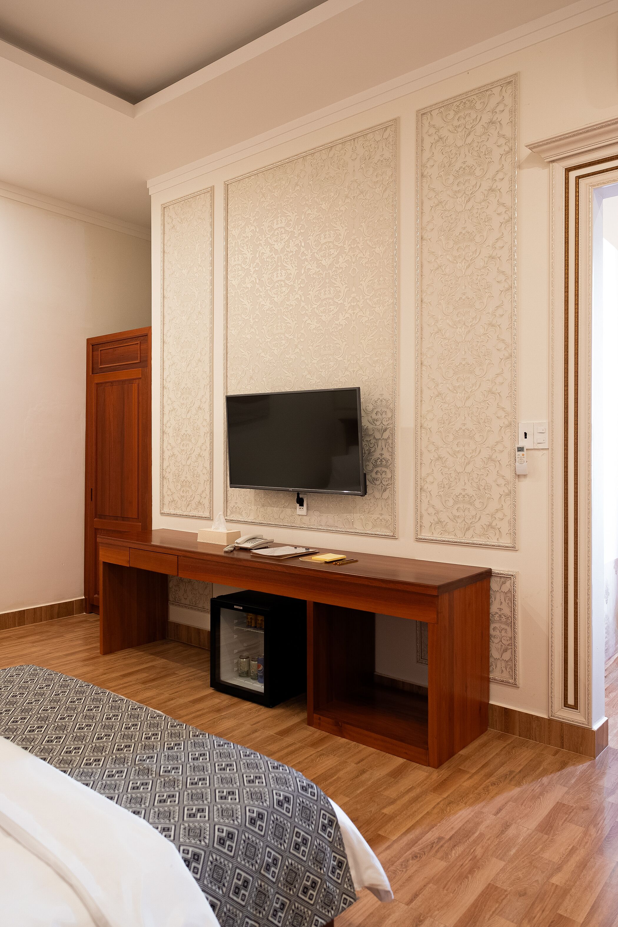 A bedroom with a wooden desk, a flat-screen TV mounted on the wall, a bed with patterned bedding, and a wardrobe.