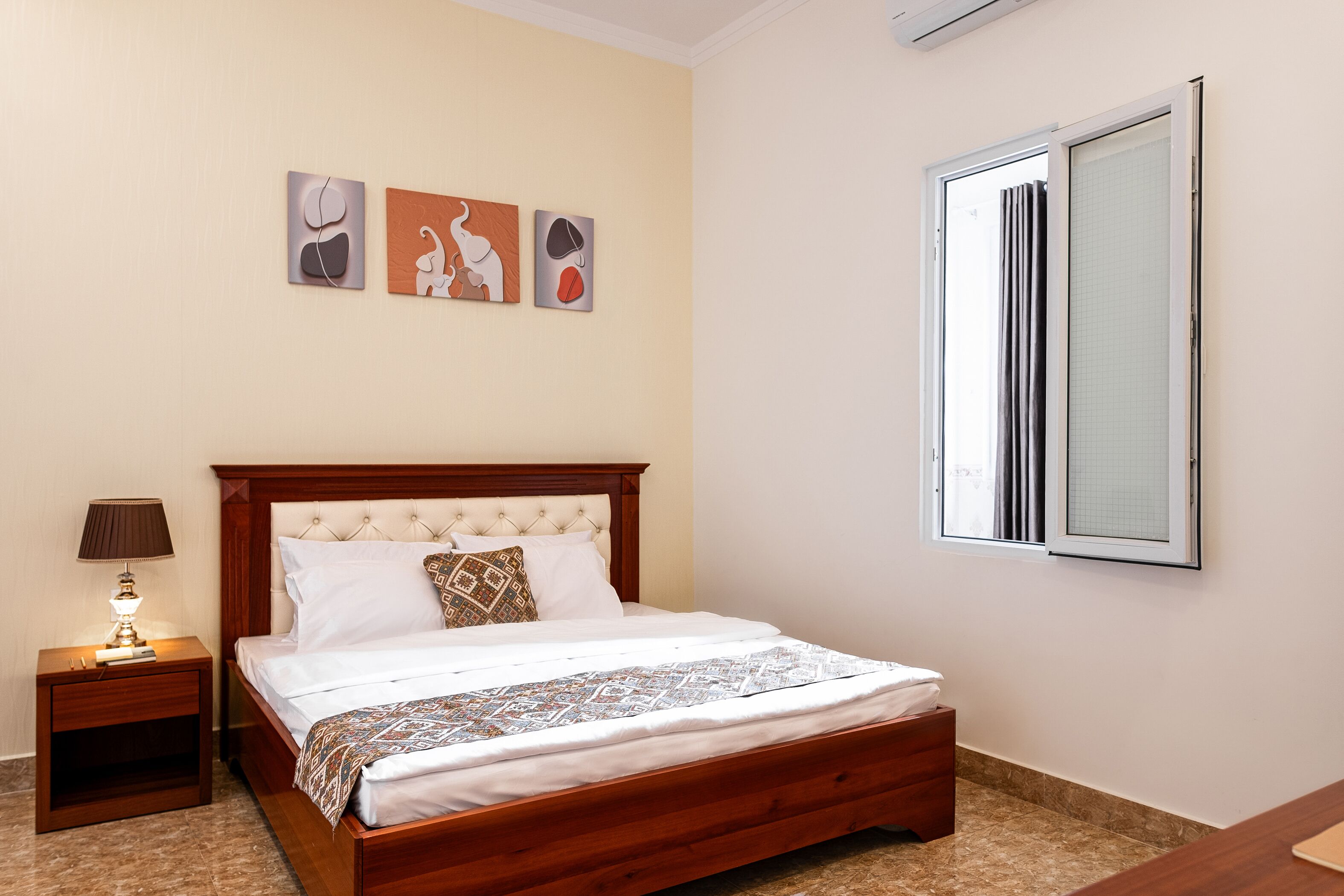 Una cama bien tendida con una colcha estampada, una mesita de noche de madera con una lámpara, y tres cuadros en la pared sobre la cama.