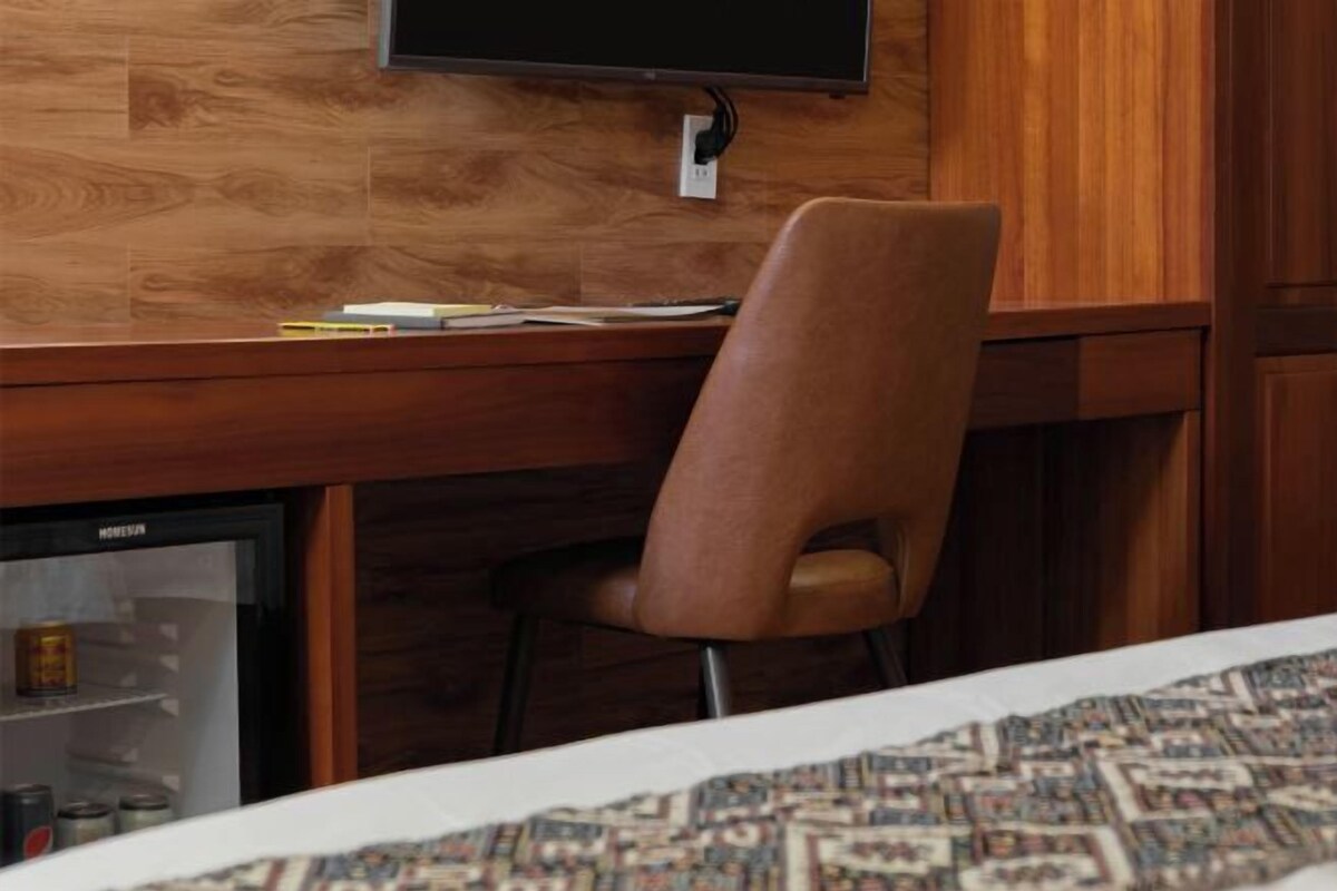 Uma mesa de madeira com monitor de computador, uma cadeira e uma geladeira com bebidas.