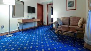 A hotel room with a blue patterned carpet, a sofa, a desk with a flat-screen TV, a mirror, and a framed picture on the wall. - City Plaza Hotel (Al-Fujairah)