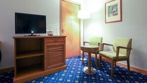 A hotel room with a TV on a wooden cabinet, a desk, two chairs, a lamp, and a framed picture on the wall. - City Plaza Hotel (Al-Fujairah)