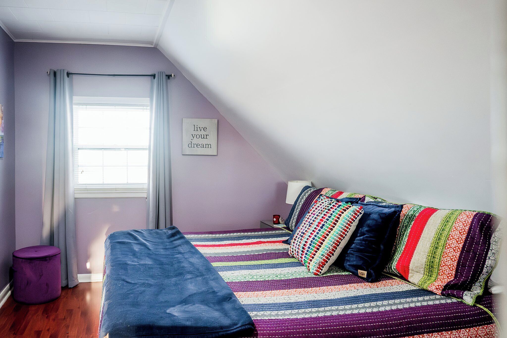 Een slaapkamer met een bed, een raam met gordijnen, een klein krukje en een motiverende quote op de muur.