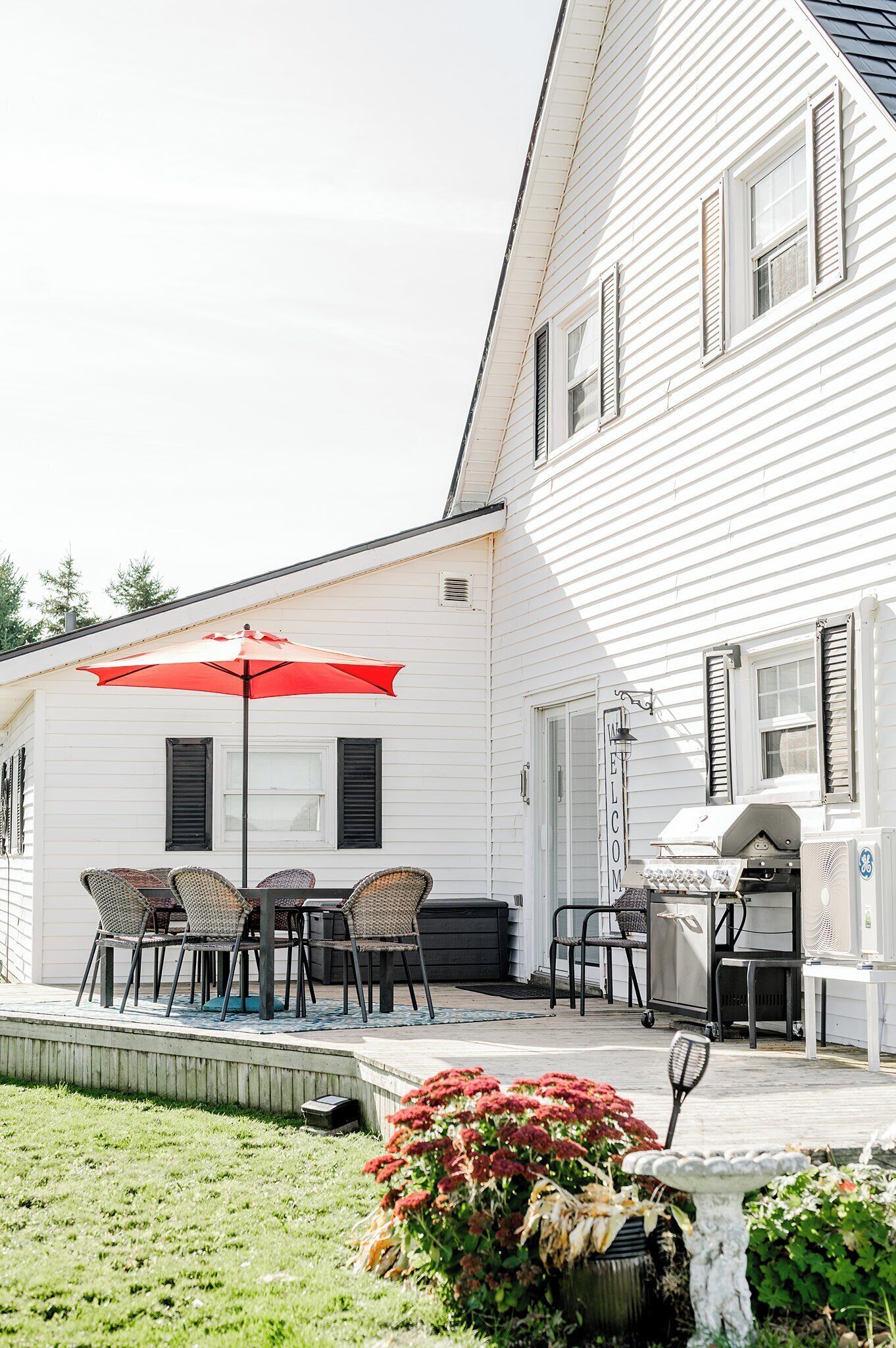 A white house with a barbecue area, outdoor seating with a red umbrella, and a welcome sign.