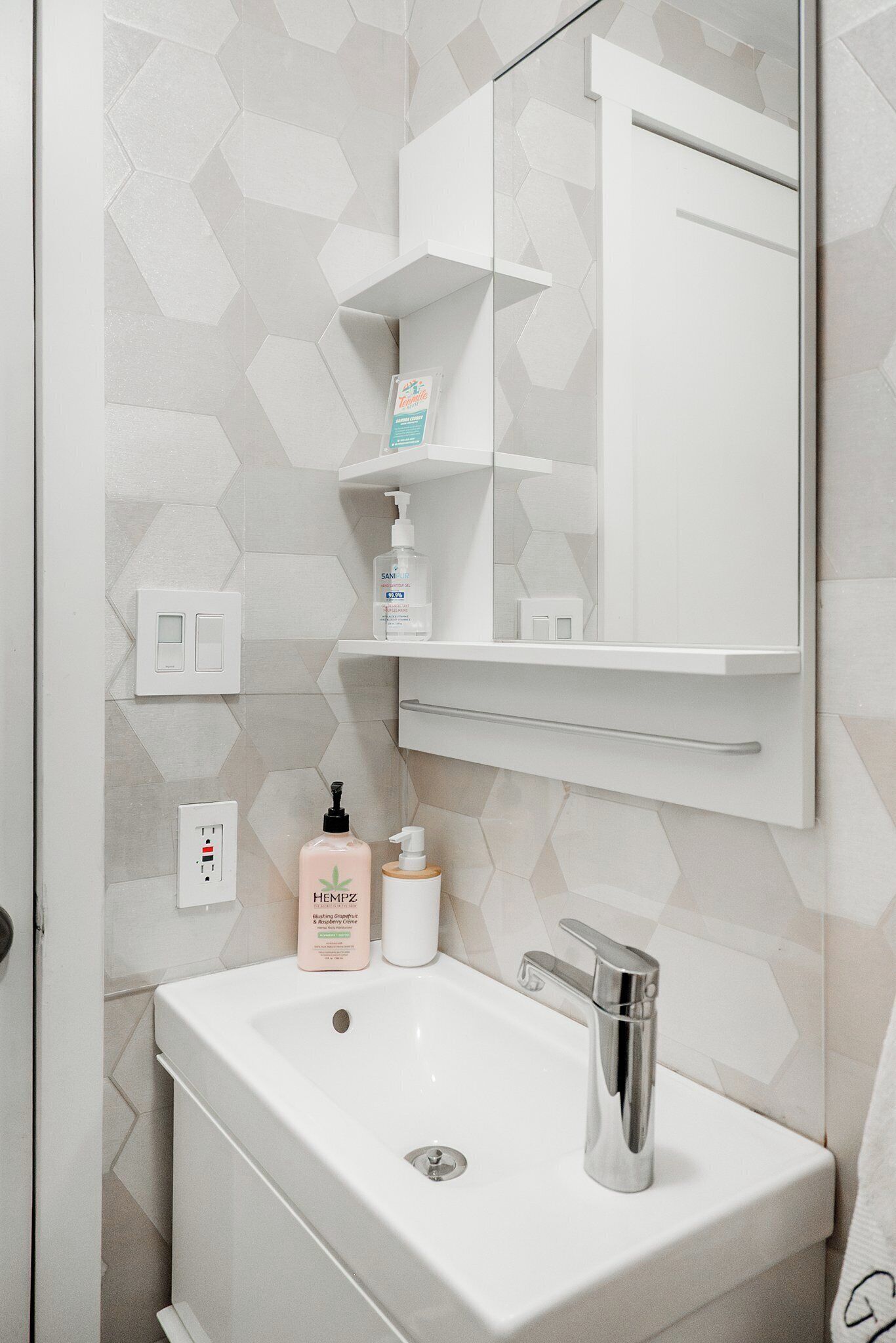 Kylpyhuone, jossa on valkoinen pesuallas, peili ja hylly, jossa on hygieniatuotteita.