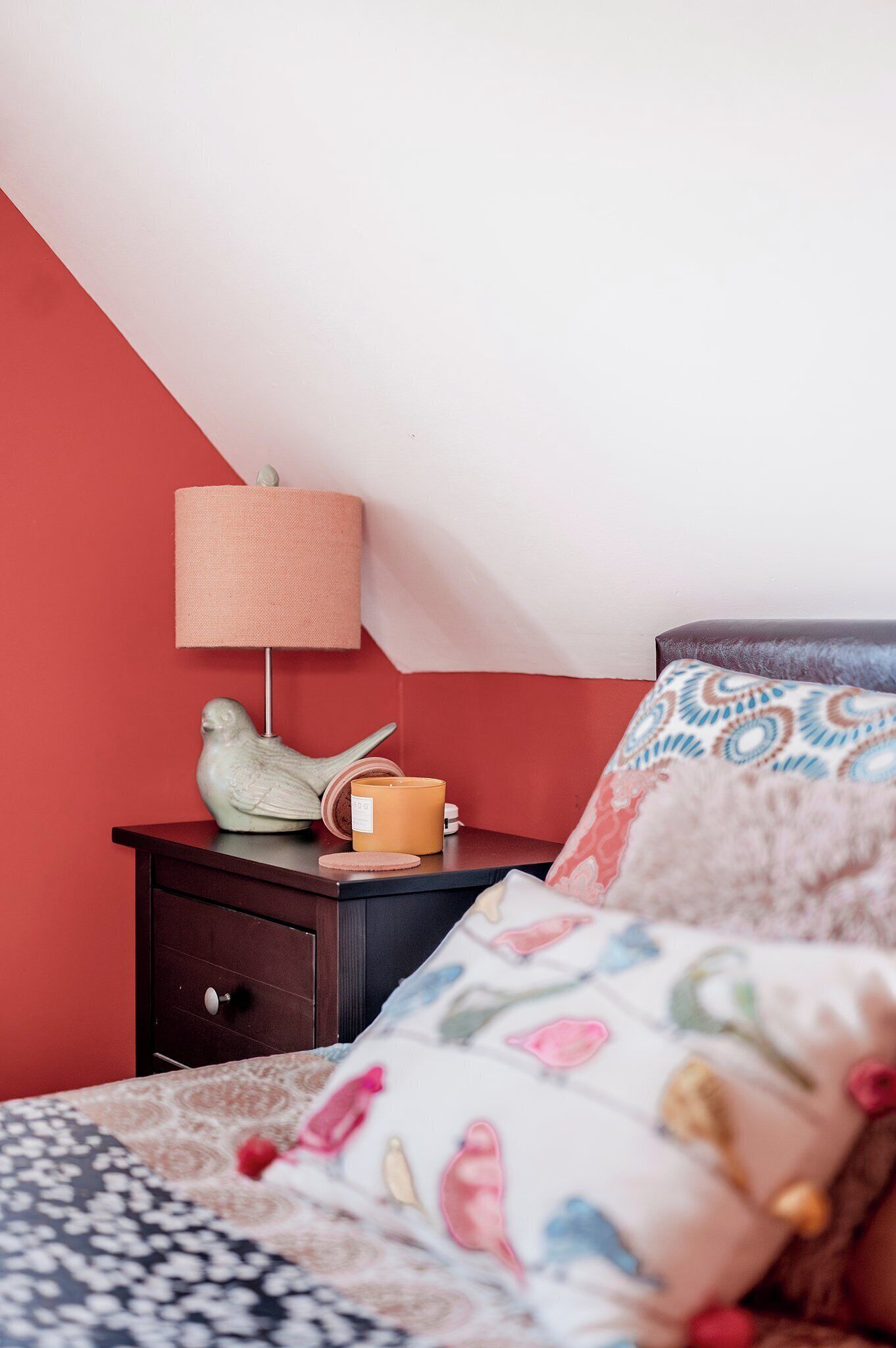Une chambre avec un mur rouge, une table de chevet avec une lampe, un objet en forme d’oiseau et un coussin à motifs.