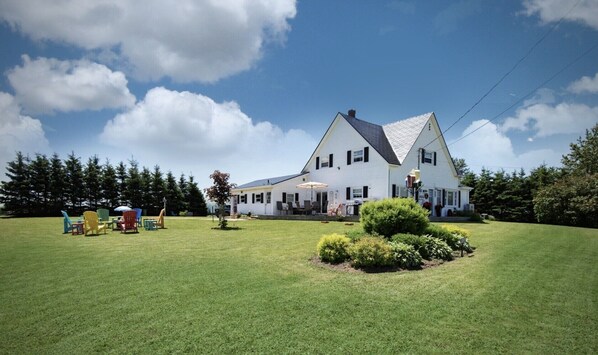 Une grande maison blanche avec un toit en pente, une pelouse bien entretenue et un espace extérieur aménagé pour s’asseoir.