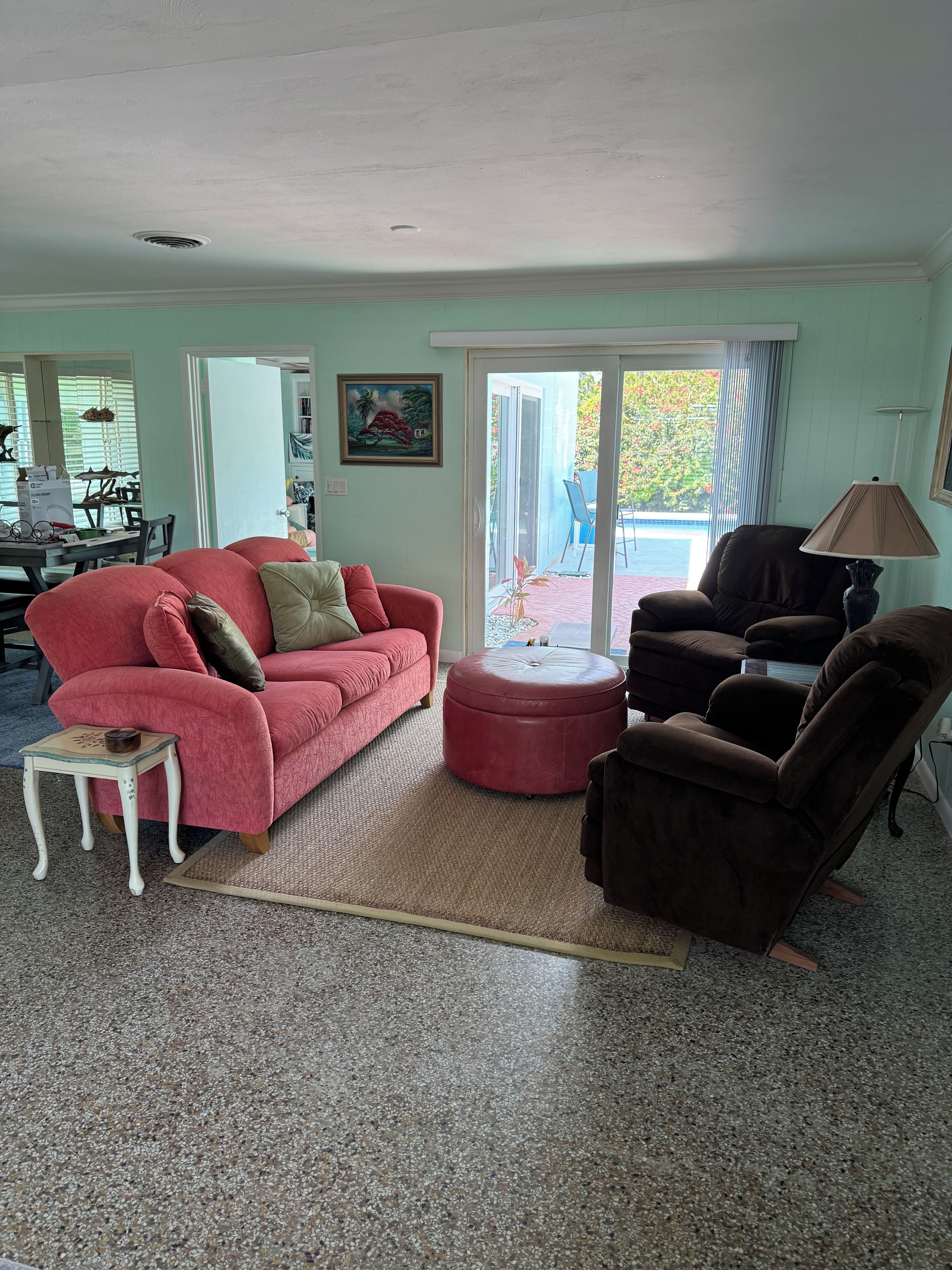 Ein Wohnzimmer mit einem roten Sofa, einem braunen Sessel, einem kleinen Tisch und einem Glastisch. Eine Tür führt zu einem Balkon mit Blick auf Grünflächen.