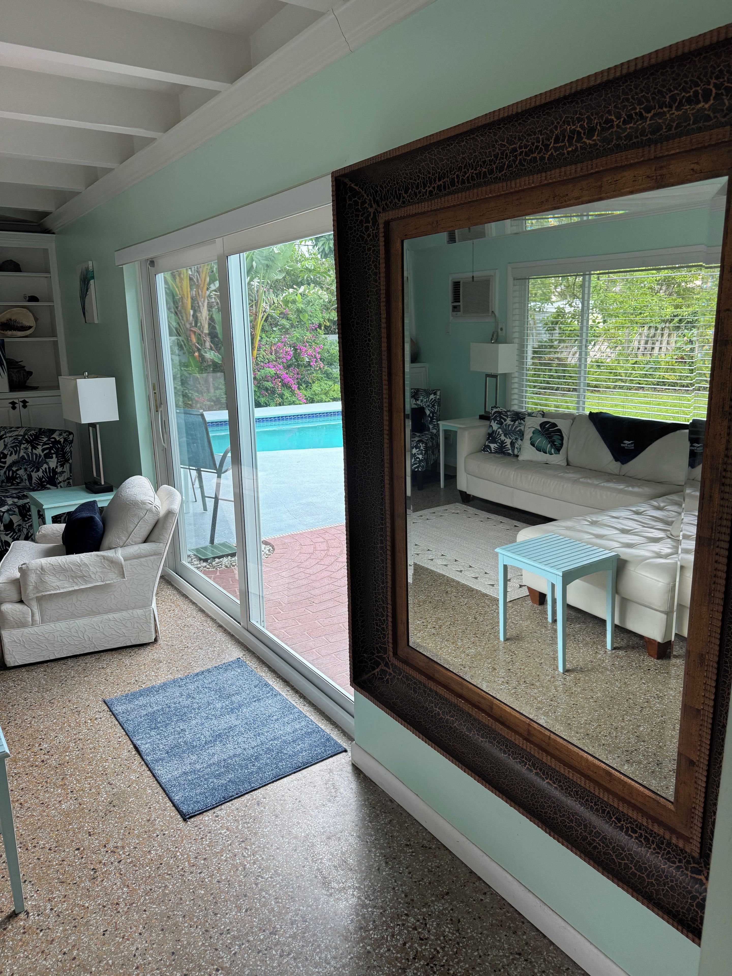 A living room with a large mirror, a sofa, a coffee table, and a view of a pool and greenery.