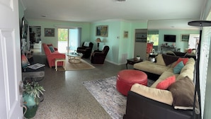 Ein großzügiges Wohnzimmer mit einer großen Eckcouch, einem roten Hocker und einem Couchtisch. Im Hintergrund ist ein Kamin, ein Fernseher und ein Essbereich zu sehen.