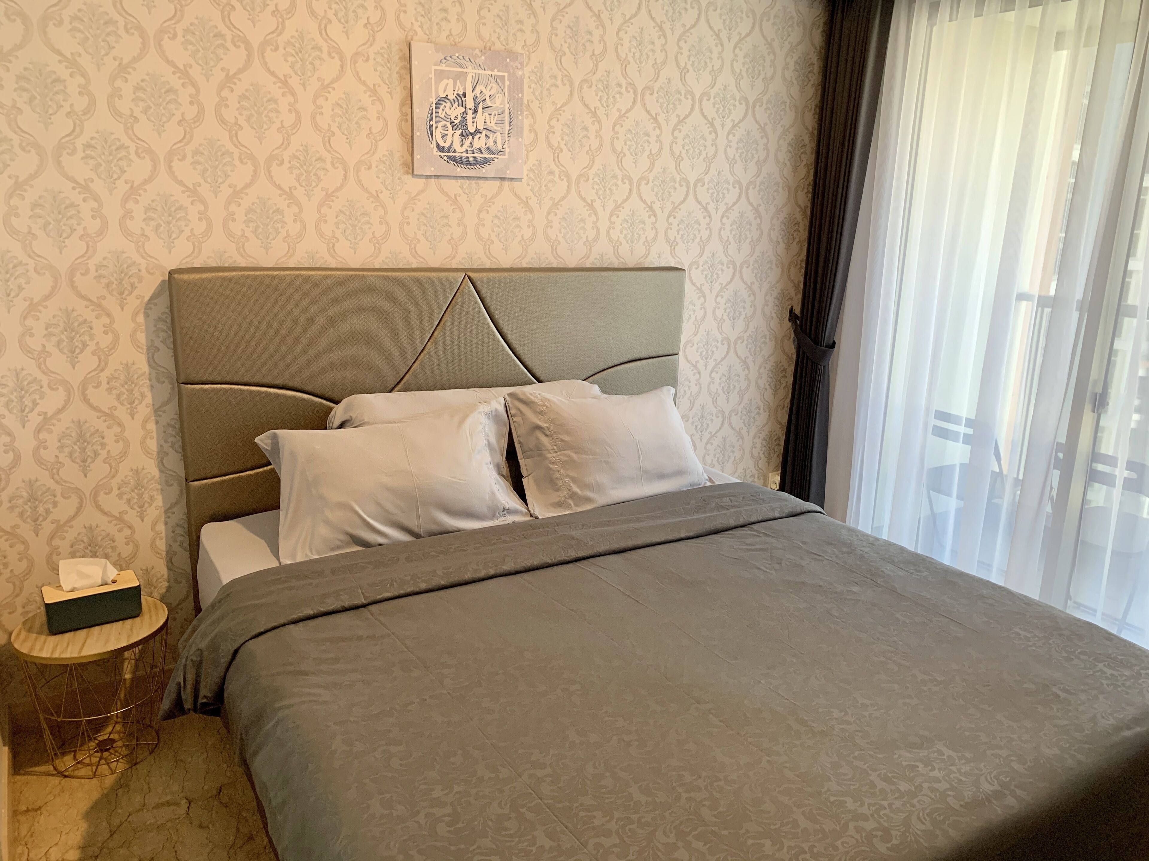 A neatly made bed with a grey bedspread and pillows, a bedside table with a tissue box, and a wall with a patterned wallpaper.