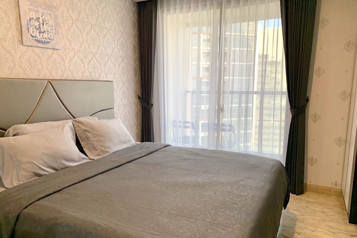A neatly made bed with a grey bedspread and pillows, a headboard, a window with sheer curtains, and a patterned wall.