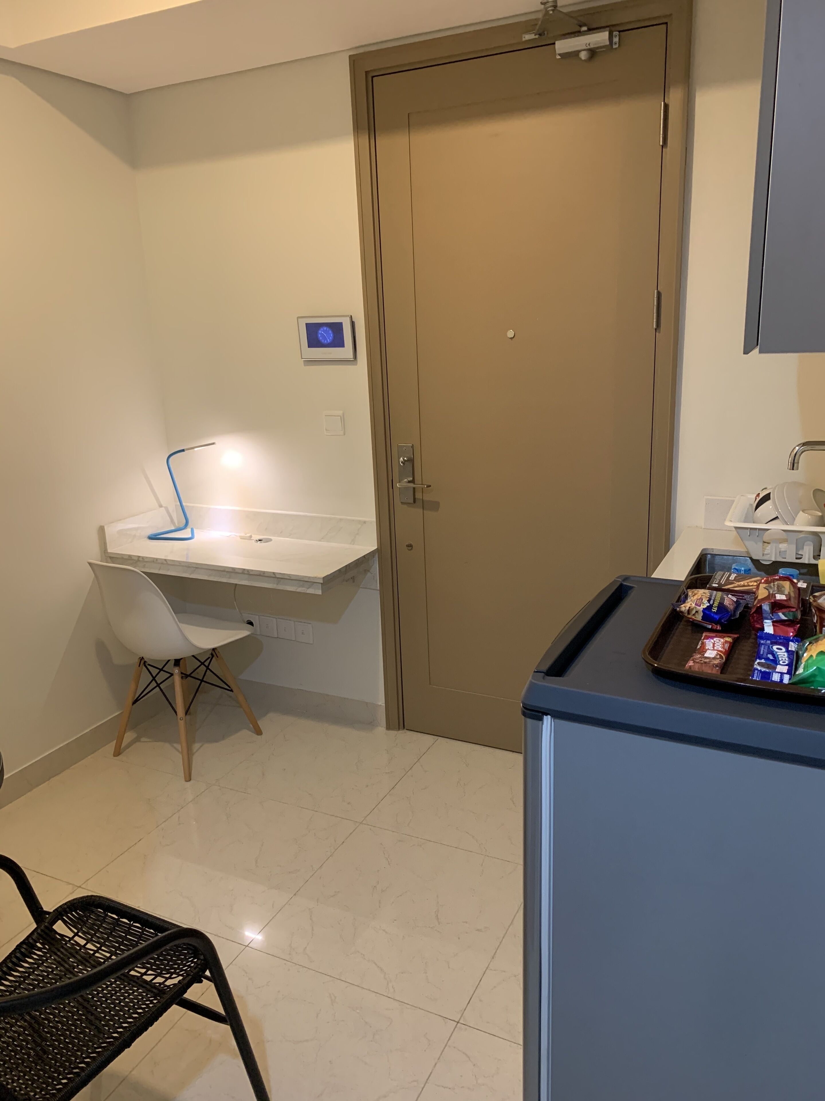 Een kleine kitchenette met een wit aanrecht, een spoelbak en een stoel. Er is een deur met een beveiligingscamera erboven, en een dienblad met snacks op een blauwe kast.