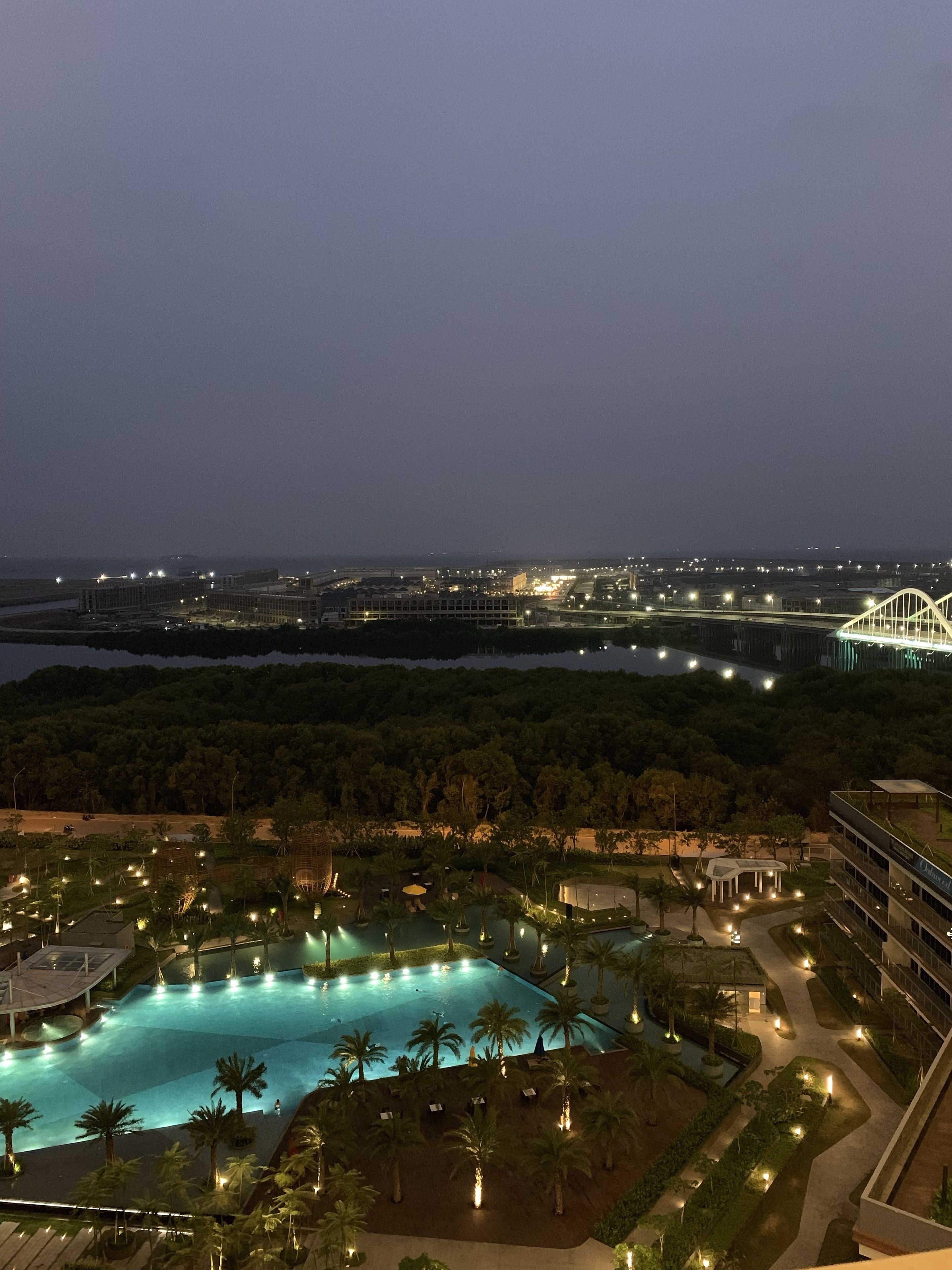 A nighttime view of a resort with a large pool surrounded by palm trees and a bridge in the background.