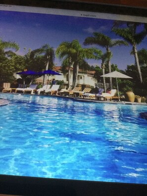Une piscine entourée de palmiers et d’un bâtiment à toit de tuiles, avec des chaises longues et des parasols à proximité.