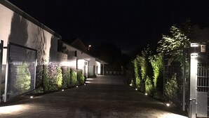 A nighttime view of a paved pathway flanked by buildings and greenery, with illuminated walls and a gated entrance. - TLA Modern and cozy holiday house freestanding (Waldmohr)