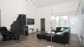 A modern living room with a black leather sofa, a black leather chair, a white coffee table, and a floor lamp. There is a staircase with a black wall and a TV mounted on the wall. - TLA Modern and cozy holiday house freestanding (Waldmohr)