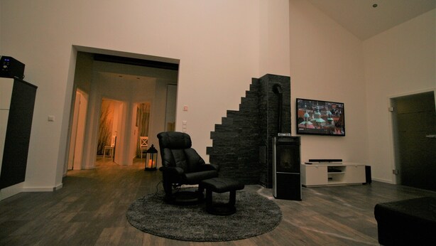 A modern living room with a dark leather chair, a flat-screen TV mounted on the wall, and a staircase leading to another level. - TLA Modern and cozy holiday house freestanding (Waldmohr)