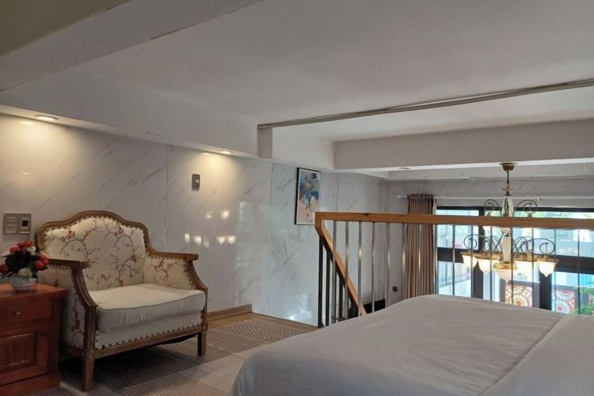 A bedroom with a large bed, a wooden chair, a dresser, and a view of the outside through a window.