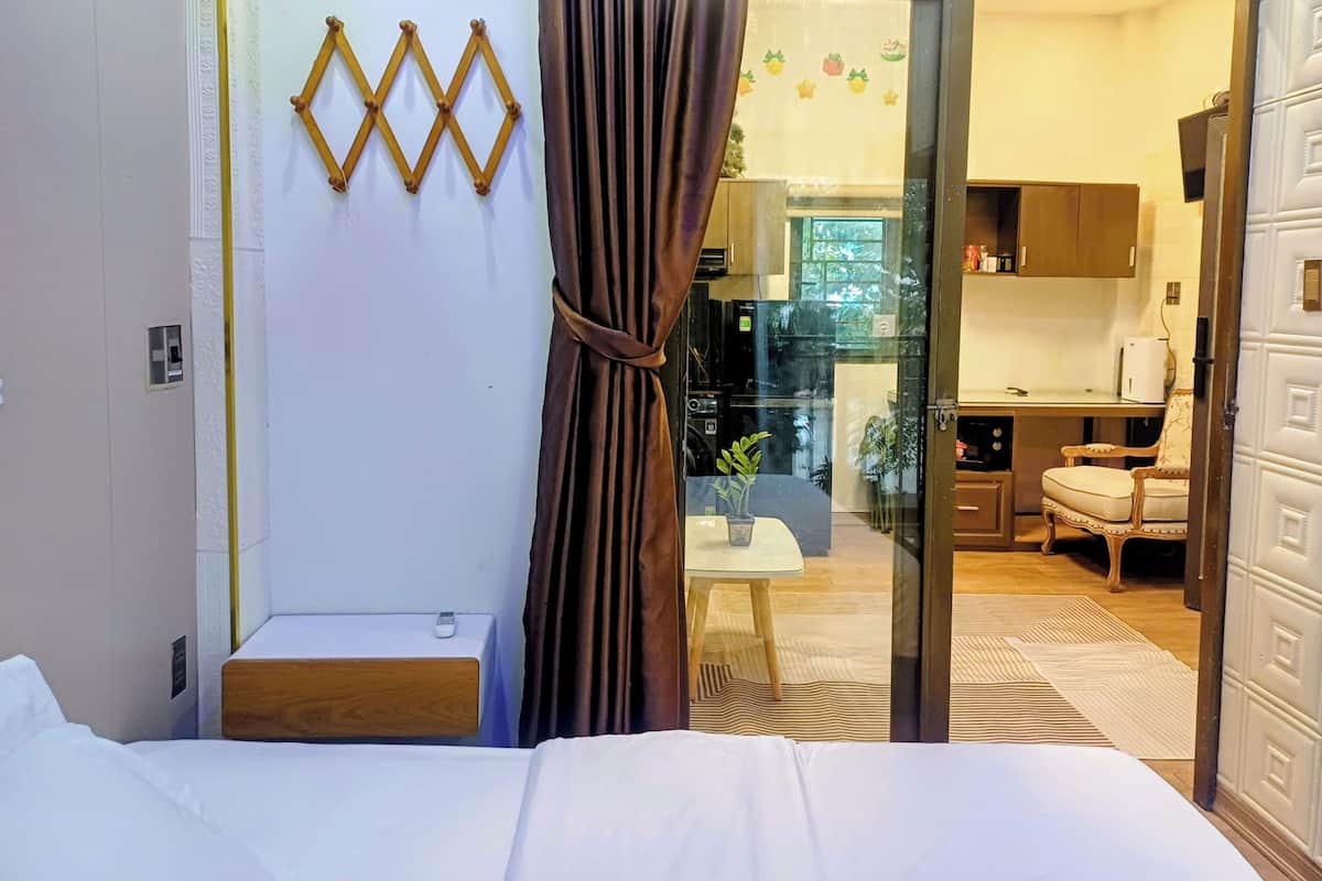 A hotel room with a bed, a small seating area, a kitchen visible through a glass door, and a decorative wall piece.