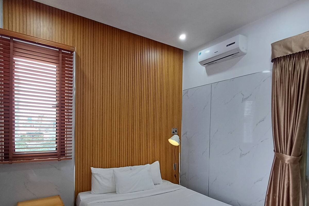 A bedroom with a wooden panel wall, a bed with white bedding, an air conditioning unit, a bedside lamp, and a window with blinds.