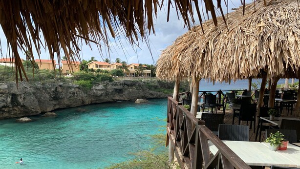Une terrasse en bois surplombant une piscine à l’eau turquoise et limpide, avec une structure de toit en chaume.