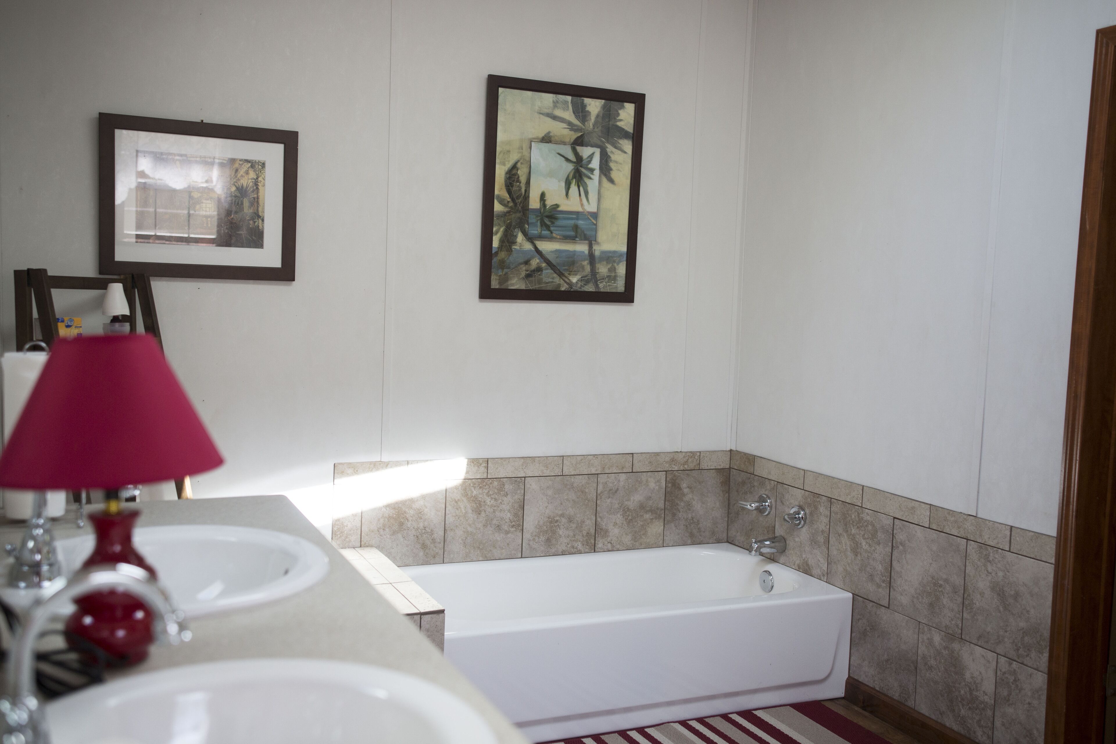 A bathroom with a bathtub, two sinks, a red lamp, and two framed pictures on the wall.