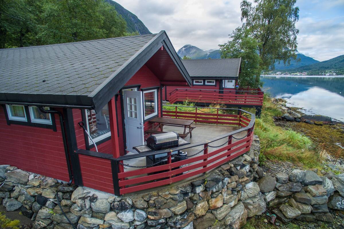 A red cabin with a stone foundation, a deck, and a view of a lake and mountains.
