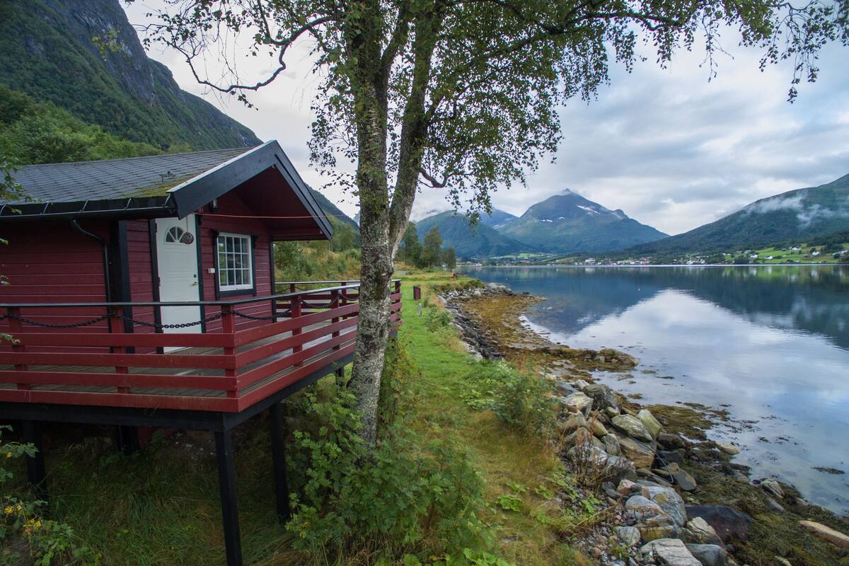 A red wooden cabin with a balcony overlooking a calm lake and mountains.