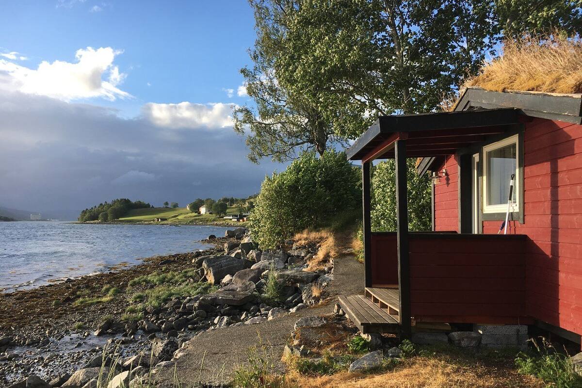 A small red cabin by a rocky shoreline with a view of the water and distant hills.