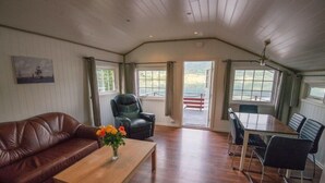 A spacious room with a wooden floor, a leather sofa, a dining table with chairs, and a view through the open door. - Fagervik Camping (Vestnes)