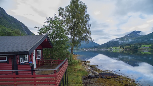 A red wooden cabin with a deck overlooking a calm lake, surrounded by mountains and greenery. - Fagervik Camping (Vestnes)