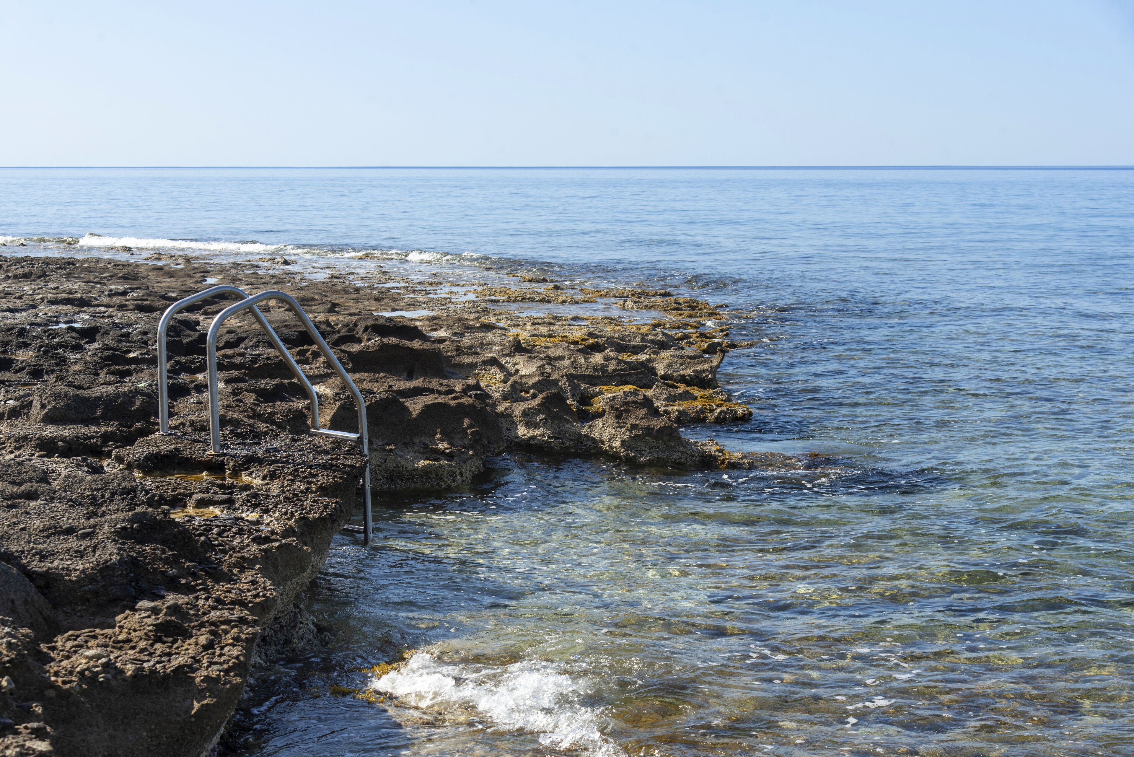 Una costa rocosa con agua cristalina y una escalera metálica que da al mar.