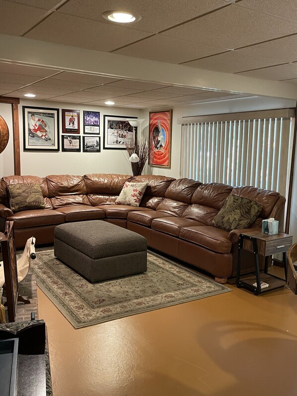 A living room with a brown leather sectional sofa, a grey ottoman, and a small side table with a tissue box. There are framed pictures on the wall, a vase with branches, and a window with blinds.