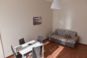 A small, well-lit room with a dining table, chairs, a sofa, and a wall-mounted lamp.