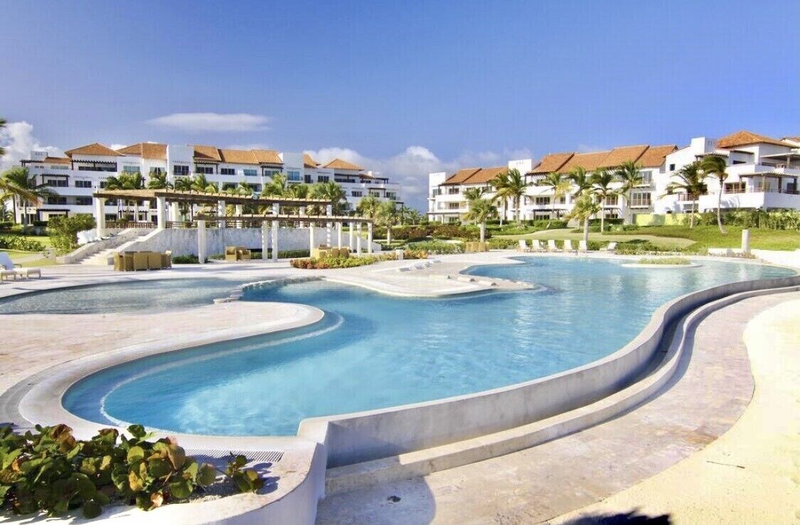 Ein Resort mit einem großen, gewundenen Swimmingpool, umgeben von Palmen und wolkenlosem, blauem Himmel.