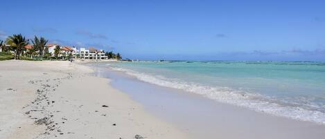 A beach with white sand, clear turquoise water, and a row of white buildings with red roofs.